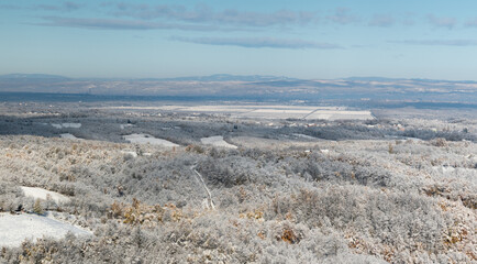 Wintry landscape of countryside, forest covered with frost and snow covered arable land, distant hills in haze