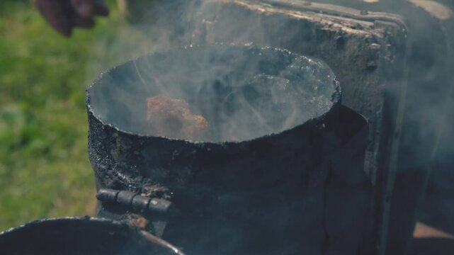 Close-up of an old beekeeper's hands lighting a smoker. Old bee smoker, beekeeping tool, held by man's hand, farm in background, concept of beekeeping 