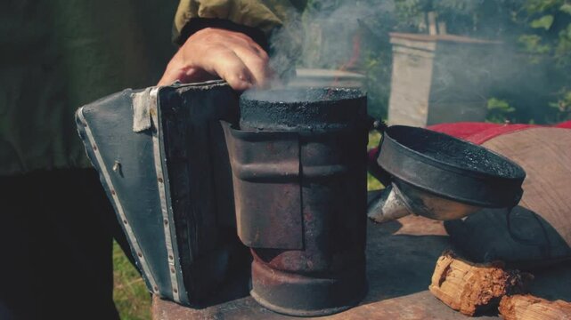 Close up of old beekeeper hands pressing the billow to light the smoker. Old smoker, beekeeping tool held by man's hand, farm in background, beekeeping concept. 