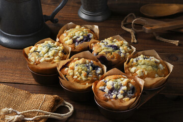Fresh baked Blueberry muffins in a muffin tin on a rustic wood kitchen table with wooden utensils and metal tea pot.