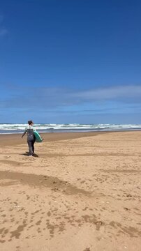 Young woman in neoprene going to water with surfboard. Female surfer on sunny day at the beach, happy to be outside and do physical activity to stay healthy. Surfing in Morocco. Vertical video.
