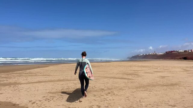 Young woman in neoprene going to water with surfboard. Female surfer on sunny day at the beach, happy to be outside and do physical activity to stay healthy. Surfing in Morocco. 