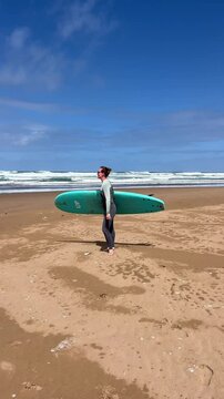 Young woman in neoprene going to water with surfboard. Female surfer on sunny day at the beach, happy to be outside and do physical activity to stay healthy. Surfing in Morocco. Vertical video.
