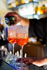 A bartender pours a vibrant orange cocktail into elegant glasses with ice on a bar counter. The background is blurred, highlighting the drink preparation in a stylish nightlife setting.