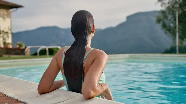 Young woman in bikini swimsuit relaxing inside swimming pool by luxury villa. Female tourist or model enjoying vacation in Italy with perfect view overlooking Tuscany mountains. Boutique hotel holiday
