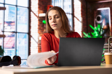 Casual person sitting at her desk and reading research papers, multitasking with laptop at home...