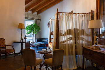 Open-space interior finely decorated with classical-style furniture, with a desk and a four-poster bed in the foreground and a sofa next to a window in the background