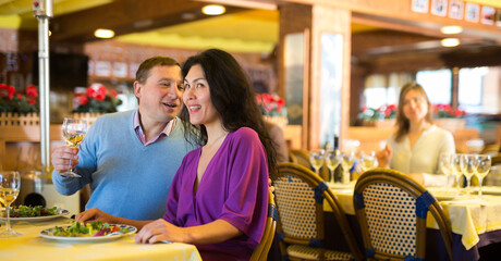 Cheerful adult interracial couple having dinner in restaurant. People sitting at served table, eating fresh vegetable salad and drinking wine