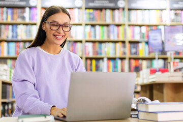 Doing research. Happy European woman in glasses using laptop computer in library, smiling at screen...