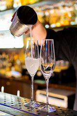 Bartender pouring a white cocktail from a shaker into two champagne glasses at a bar. The blurred background emphasizes the drink preparation process, highlighting the elegance and precision of mixolo