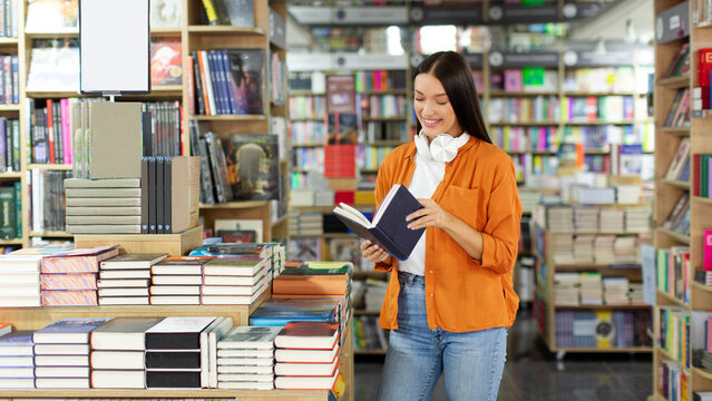 Young woman buying books at bookstore and reading one while choosing best seller, bookshelves on background