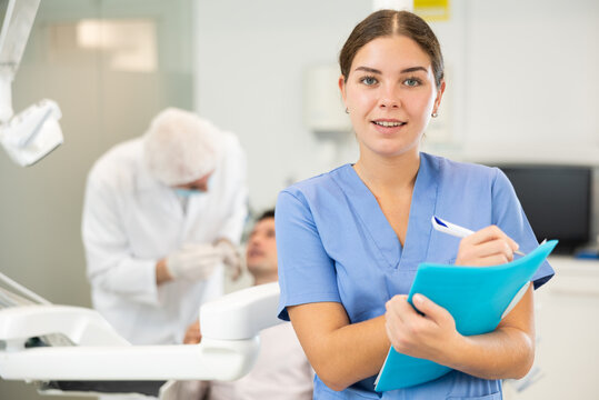 Young woman nurse in blue suit uniform with paper is standing in dental office, clinic employee in suit uniform near dentists workplace. Doctor enjoys working in profession.