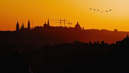 Fototapeta premium Silhouette of city skyline at sunset. Construction cranes are visible. Birds in flight. Warm orange hues. Silhouettes of buildings and rooftops