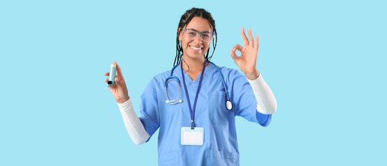 Young African-American female doctor with stethoscope and asthma inhaler showing OK gesture on blue...