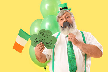 Mature man with paper mustache, card and Irish flag on yellow background. St. Patrick's Day...