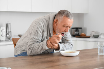 Senior man suffering from Parkinson syndrome eating soup at table in kitchen