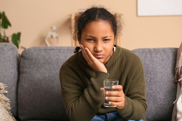 Cute African-American girl with glass of water suffering from tooth ache on sofa at home
