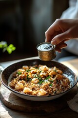 Hand seasoning a bowl of cooked shrimp and grain dish with fresh herbs in natural light on rustic wooden surface