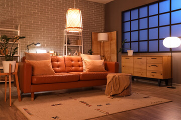 Interior of modern living room with brown sofa, chest of drawers, shelving units and glowing lamps at evening