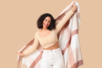 Beautiful young African-American woman with cozy blanket on beige background