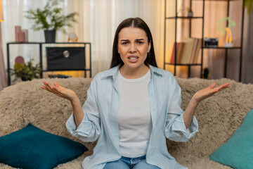 Young confused woman at home on sofa making surprised What Why facial expression looking shocked,...