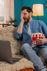 Adult man at home enjoys popcorn while watching movie on laptop, fully immersed in scene. Caucasian...