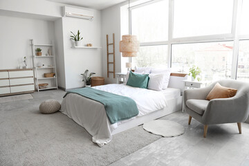 Interior of light bedroom with cozy bed and grey armchair near big window