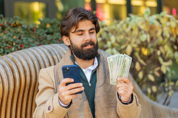 Happy rich Indian businessman tourist using smartphone and counting money dollar cash banknotes...