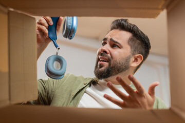 Dissatisfied annoyed male customer open cardboard box receive damaged broken wireless headphones,...