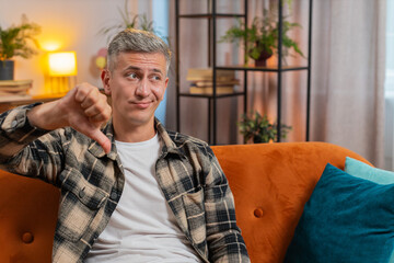 Fototapeta premium Dislike. Upset unhappy young Caucasian man showing thumbs down sign gesture, expressing discontent, disapproval dissatisfied bad work at home apartment indoors. Displeased guy with gray hair on sofa.