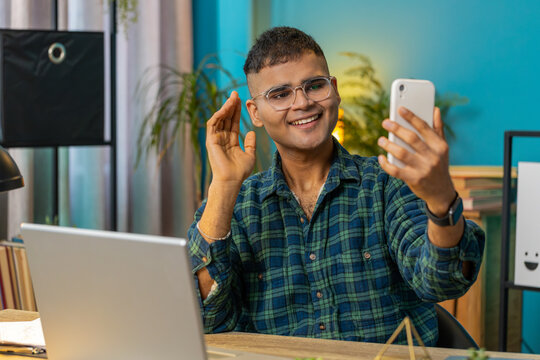 Happy Indian man blogger taking selfie on smartphone communicating video call online with subscribers recording stories for social media. Hispanic guy sitting at home office table workplace. Lifestyle