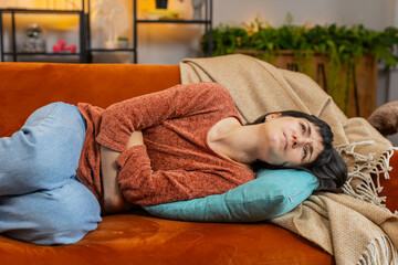 Obraz premium Upset young woman embracing belly suffering from stomachache lying on couch. Unhappy Caucasian girl having menstrual painful feelings, resting on couch. Gastritis, abdominal or period pain concept