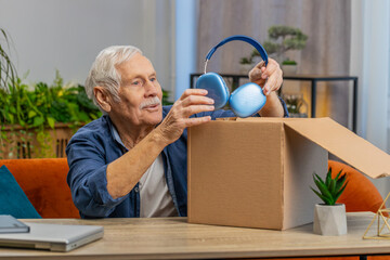 Happy senior man unpacking delivery parcel with headphones at home. Smiling satisfied grandfather...