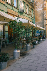 Obraz premium Outdoor café with cream awnings, metal pots with lush plants, and classic European architecture, creating a cozy and inviting street dining atmosphere