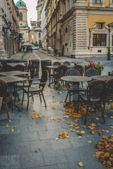 Empty outdoor café with wooden chairs and tables on a stone street, covered with fallen autumn leaves in a historic European city center