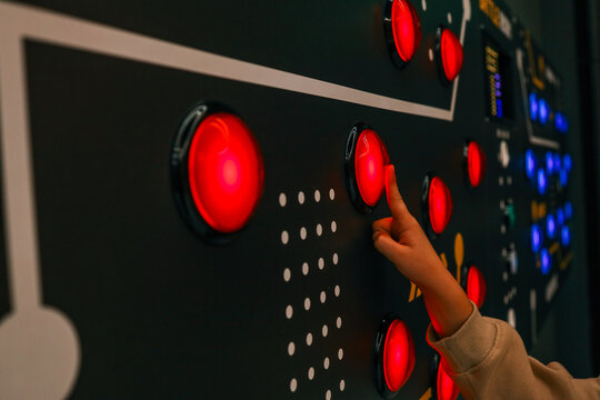 Child’s hand pressing a glowing red button on an interactive control panel with multiple lights, in a modern science or technology exhibit