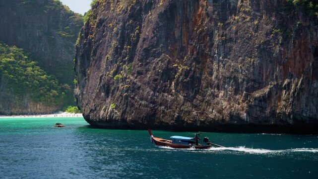 Turquoise waves washing rocky shoreline, longtail boat gliding past maya bay, phi phi leh island, trailing white wake under soft sunlight