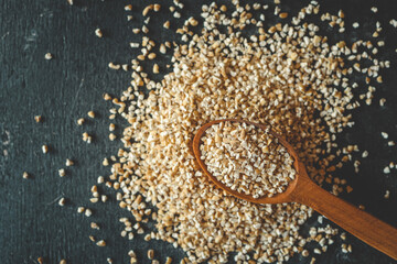 Close-up of raw steel-cut oats in a wooden spoon with scattered grains on a dark surface. Rustic...