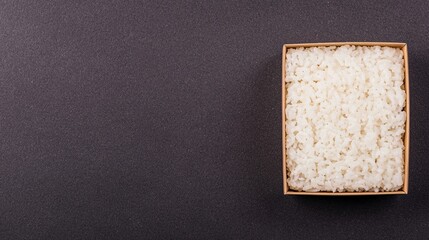 A square wooden box filled with cooked white rice sits on a dark textured surface, positioned in the upper right corner.