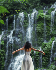 Back View of Woman in White Dress with Arms Outstretched at Bali Waterfall