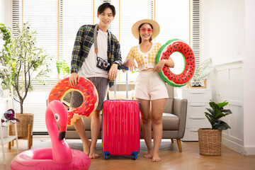 Young Asian couple holding a camera and preparing for travel in a cozy living room, with a sofa, rubber ring, luggage, and sunglasses, summer time concept.