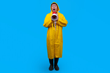 Full length of shocked man in raincoat yelling in megaphone isolated on blue background © Gatot