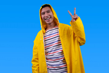 Asian man wearing yellow waterproof raincoat showing peace sign isolated over blue background © Gatot