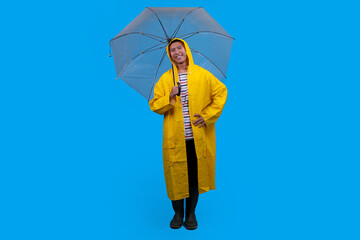 Young Asian male stands confidently with umbrella, dressed in waterproof raincoat over blue background. © Gatot