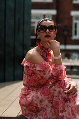 Fashionable woman wearing sunglasses and pink floral dress posing outdoors
