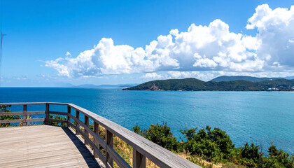 Obraz premium stunning view from wooden observation deck overlooking serene blue ocean, with lush green hills and fluffy white clouds in background, creating peaceful atmosphere