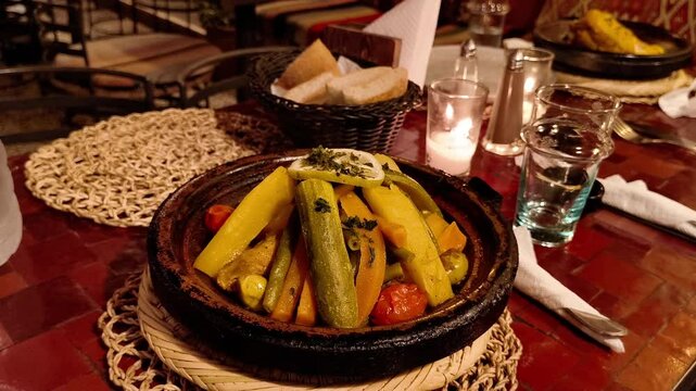 A tagine, filled with colorful cooked vegetables and garnished with lemon and herbs, sits on a straw trivet on a red patterned table. Bread in a basket, drinking glasses, a lit candle, are visible.