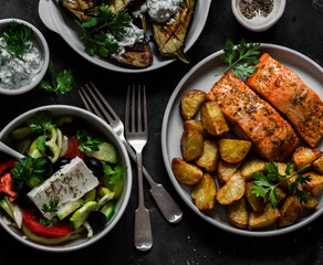 A varied lunch - baked salmon with potatoes, Greek salad, grilled eggplant with Greek yogurt sauce on a dark background, top view