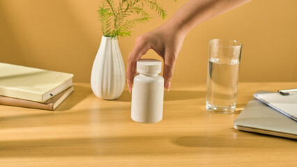 Front view of a white unlabeled pill bottle, a hand is holding the bottle from above and holding it in mid-air. Surrounding it are work items such as a laptop, prescription, and notebooks.