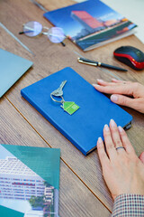 Close-up of a real estate workspace with a blue notebook, a house keychain, brochures, and a laptop on a wooden table. 
